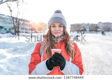 Close-up of a girl in winter outside. In warm clothes, in the hands of a mug with a hot drink of coffee tea. Emotions of joy fun and outdoor recreation. Long hair a warm winter jacket and hat.