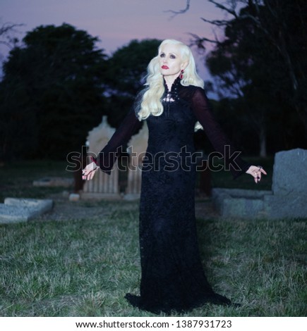 Photo of a pretty blonde woman wearing a long black dress standing in an old graveyard site during autumn twilight in the evening  