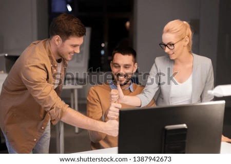 business, success and teamwork concept - happy coworkers with computer working late at night office making thumbs up gesture