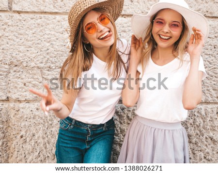 Two young beautiful blond smiling hipster girls in trendy summer white t-shirt clothes. Women posing in the street near wall. Positive models having fun in sunglasses and hat. Showing peace sign