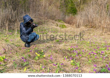 A girl in a warm jacket in nature taking pictures of flowers in a clearing