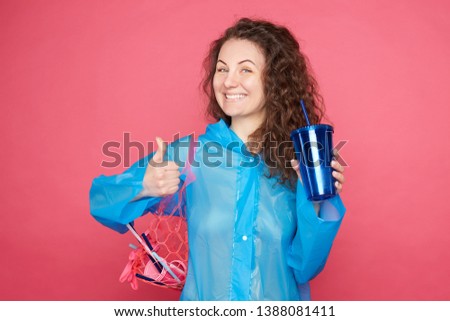 Satisfied curly haired woman in good mood, cares about environment, carries bag with plastic wastes, has rubbish stuck in curly air, participates in volunteer activities, posing on pink background.