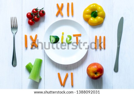food clock with Roman numerals. white plate with the inscription "diet" of vegetables, spoon and fork.Celery, Apple, yellow pepper, cherry tomatoes on a plate. on white wooden background
