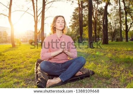 Beautiful young woman sitting in a pose of a lotus on city park sunset background. Practice meditation, kundalini yoga. Female eyes closed and open energy chakras. 