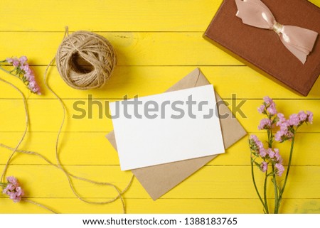 Flat lay, top view office table desk. Feminine desk workspace with blank paper card, craft envelope, violet flowers, gift box, twine, ribbon on yellow wooden background. Home office desk concept.