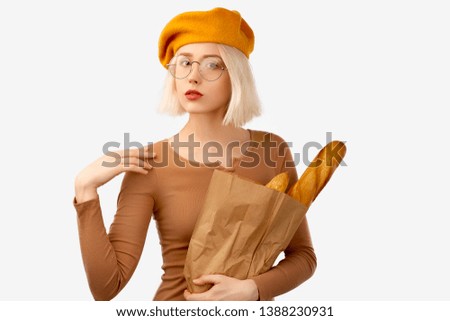 Young woman holding a bag with baguette. Serious female traveller keeps arm on shoulder, looks self assured, dressed in brown shirt, yellow beret, large round glasses, ready to explore unknown city.