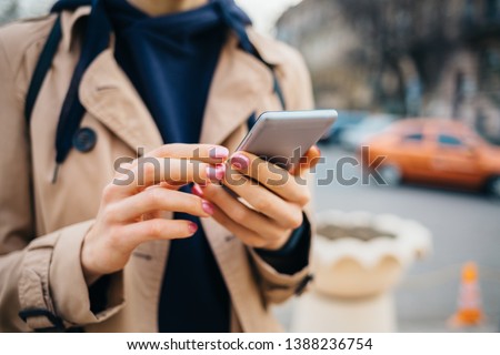 Young woman wearing beige coat using smart phone standing near road with cars in city at spring day. Close-up female's hands holding mobile phone outdoors. Royalty-Free Stock Photo #1388236754