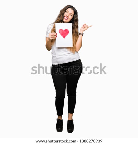 Young adult woman holding card with red heart very happy pointing with hand and finger to the side