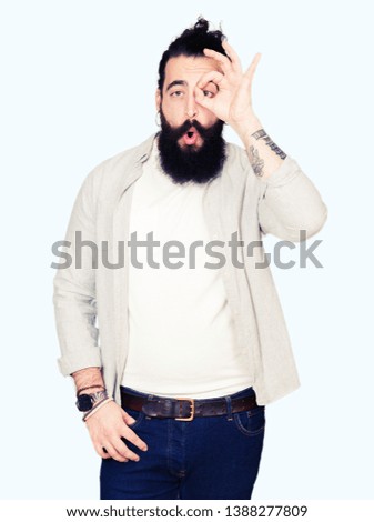 Young man with long hair, beard and earrings doing ok gesture shocked with surprised face, eye looking through fingers. Unbelieving expression.