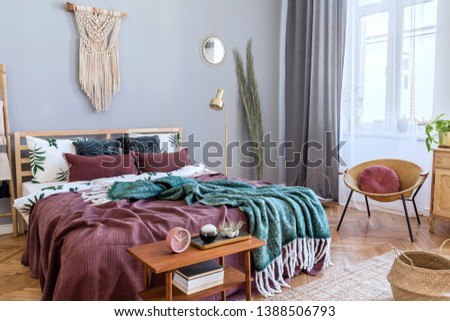 Stylish and luxury interior of bedroom with design furnitures, honey yellow armchair, gray macrame, gold lamp and elegant accessories. Beautiful bed sheets, blankets and pillows. Cozy home decor.