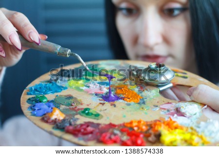 Closeup of a young woman with black hair mixes paint on a palette with a spatula against a white canvas, creating an oil painting, soft focus