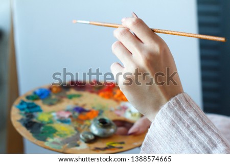Close-up of female hands in a pink sweater holding a brush and a palette with multi-colored acrylic paints against a white canvas, selective focus