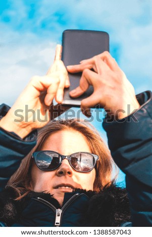 Young woman with brown hair wearing sunglasses with a black smartphone taking a selfie outdoor – Girl holding the phone up and looking at it – Concept image for teens using gadgets