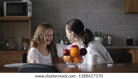 Happy mother and daughter sit in the kitchen at the table and have fun talking. An adult woman and a schoolgirl laugh and look at each other.