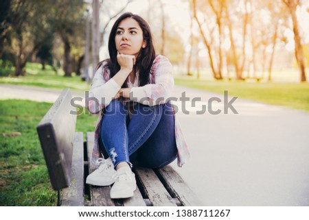 Serious attractive girl thinking about plans in park. Or she waiting for friend. Pretty woman sitting on bench and looking away. Contemplating concept