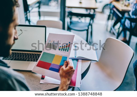 Cropped view of young man holding paper document with colorful diagrams of financial reports and checking information working remotely at modern laptop computer connected to wireless in coworking