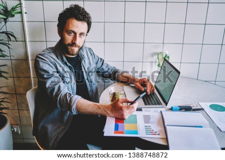 Portrait of bearded young man holding smartphone in hand and looking at camera during remote work at laptop in coworking.Hipster guy chatting on phone sitting at desktop with paper diagrams