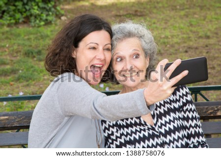 Two joyful ladies having fun and grimacing for selfies in city park. Senior woman and her adult daughter enjoying weekend outdoors. Happy family concept