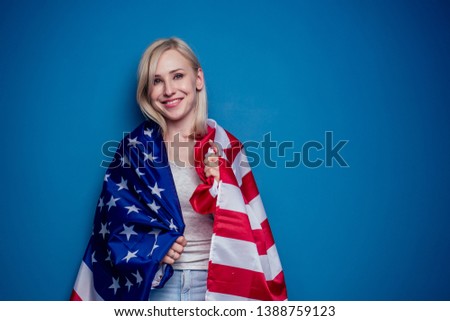 blonde woman in jeans holding American flag with paper crown and torch Statue of liberty on a blue background in the studio .4th july independence day celebration concept