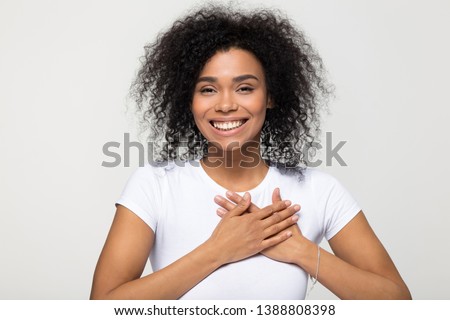 Head shot portrait happy african woman in white t-shirt pose grey background smiling looking at camera holding hands on chest feels gratitude, gesture of sincere feelings from heart and love concept Royalty-Free Stock Photo #1388808398