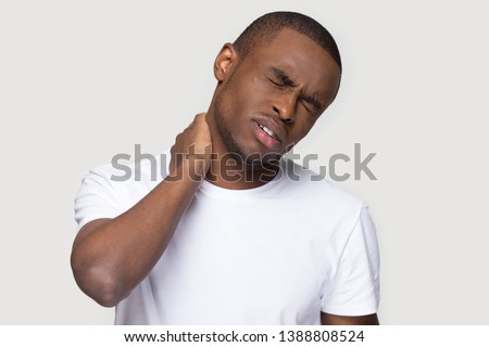 Black millennial man in white t-shirt on grey black background guy feels physical discomfort unhealthy tired closed his eyes for neck pain massaging tensed muscles relieve joint shoulder ache concept Royalty-Free Stock Photo #1388808524