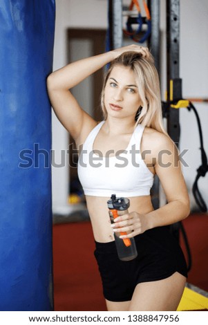 Young pretty blond woman standing in a gym near a boxing pear and holding a bottle of water in her hands. Active Lifestyle. Sports in the gym.