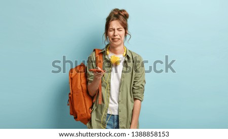 Miserable dejected hipster girl cries and sobs from despair, holds modern smart phone, has problem, cannot browse music app, dressed in casual shirt and jeans, carries rucksack on one shoulder Royalty-Free Stock Photo #1388858153