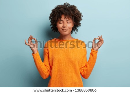 Mindful peaceful Afro American woman meditates indoor, keeps hands in mudra gesture, has eyes closed, tries to relax after long hours of working, holds fingers in yoga sign, isolated on blue wall Royalty-Free Stock Photo #1388859806