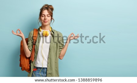 Photo of relaxed student keeps hands in mudra gesture, keeps eyes closed, listens peaceful music, headphones on neck, wears shirt and jeans, has rucksack, isolated on blue background with free space Royalty-Free Stock Photo #1388859896