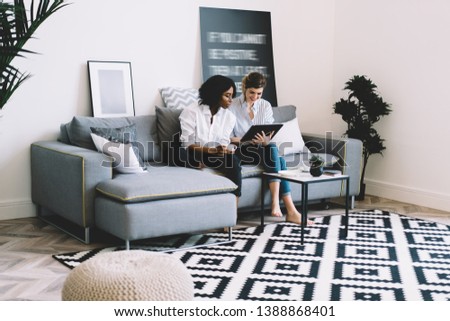 Positive caucasian young woman holding modern touch pad and showing funny photos in internet profile african american hipster girl sitting on comfortable couch in apartment with fashionable interior