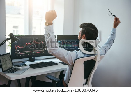 Yes! Back view of stock trader with raised hands looking at multiple computer screens with data and charts and feeling happy while sitting in modern office Royalty-Free Stock Photo #1388870345