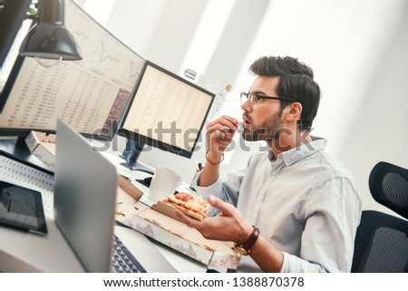 It can not be truth! Disappointed young trader is looking at monitor screen with trading charts and financial data while eating hot pizza in his modern office.