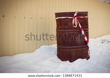 Old metal oil barrel standing in the snow with a ribbon. Steel drum is rusty, scratched, dented. Winter grunge style photography for ecology problem theme. 