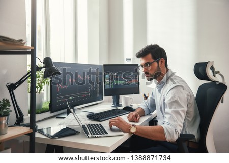 Busy working day. Young bearded trader in eyeglasses working with laptop while sitting in his modern office in front of computer screens with trading charts. Royalty-Free Stock Photo #1388871734