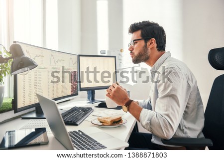 Hungry businessman. Side view of young bearded trader is eating fresh sandwich while looking at monitor screen with financial data in his modern office