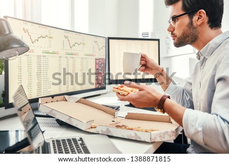 Food for productive work. Side view of young bearded trader in eyewear is eating hot pizza while looking at monitor screen with trading charts and financial data in his modern office.
