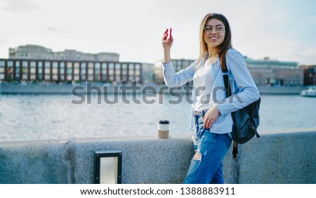 Smiling young woman tourist in stylish casual wear looking away while making photo on modern smartphone device standing in urban setting during trip.Positive hipster girl with backpack taking picture