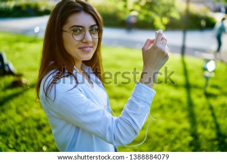 Portrait of cheerful beautiful young woman in eyeglasses smiling at camera while making photo on smartphone device enjoying music audio in modern earphones during walk in park in urban setting