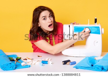 Photo of young lady works in her sewing space, has problems with sewing machine, creates accessories, repairing clothes, embroider and packs trendy clothes, sitting against yellow studio wall.