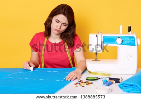 Image of attractive female fashion designer working in her workshop, being in process of creating new clothes collection, tired of long hours work, draws with soap on blue cloth, over yellow wall.