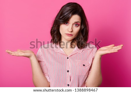 Studio shot of attractive woman shrugging helpless with her hands, wearing stylish stripped shirt, looking at camera with sad facial expression, has problems, posing against pink studio background. Royalty-Free Stock Photo #1388898467