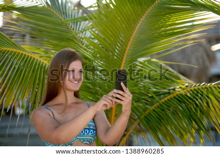 Happy young woman wearing bikini taking selfie using smartphone in the summer day between palm on the beach in Florida, USA