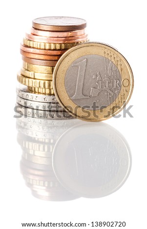 A standing 1 Euro coin - currency of European Union - leaning on the stack of euros and euro cents on white reflective surface