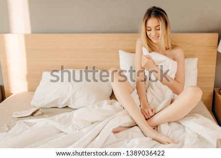 Blonde young woman with smiling beautiful face having fun and hugging pillow sitting on bed in her bedroom