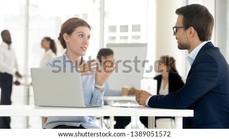 Concentrated man and woman workers sit at office desk negotiating using laptop discussing ideas, diverse colleagues talk brainstorming negotiate on business project together. Cooperation concept Royalty-Free Stock Photo #1389051572