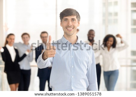 Happy Caucasian man employee or leader stand front show thumbs up recommending company service, smiling male client look at camera give recommendation, excited team support motivate at background