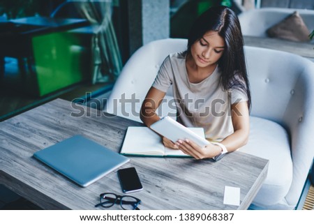 Concentrated brunette woman noting information in notebook using portable pc for research, skilled pensive female freelancer writing article for publication in blog doing remote work in cafe
