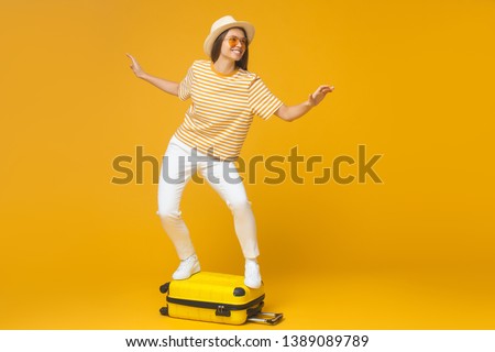 Young tourist girl standing on suitcase, pretending like she is surfing, isolated on yellow background. Dream about traveling concept