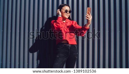 Attractive female teengaer in trendy sunglasses taking selfie pictures during leisure time near copy space for advertising text, beautiful chinese hipster girl photographing on smartphone front camera