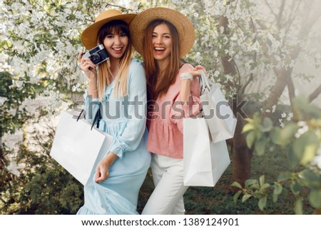 Shopaholics on sale. Summer shopping concept. Outdoor image of two  happy  tourist women in elegant dress holding white shopping bags and posing in amazing blooming garden.
